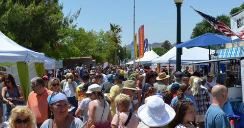 A bustling outdoor festival with crowds of people, colorful tents, and trees under a clear blue sky.