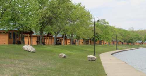 A row of wooden cabins lined along a grassy path beside a calm lake, surrounded by lush green trees.