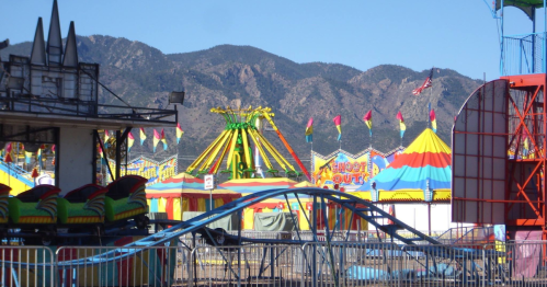A colorful carnival scene with rides, tents, and mountains in the background under a clear blue sky.