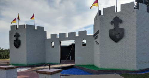 A white castle structure with flags, featuring a drawbridge and coat of arms on the towers against a cloudy sky.
