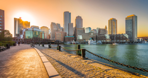 Sunset over a city skyline with skyscrapers, reflecting on water, along a cobblestone waterfront path.