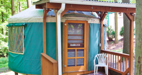 A cozy yurt with a wooden deck, surrounded by trees, featuring a door and a white chair outside.