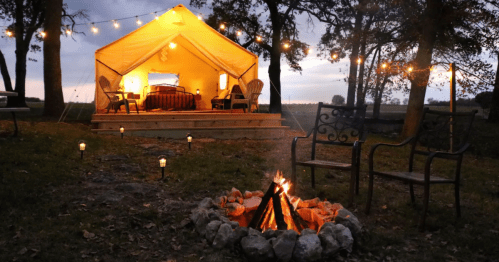 A cozy glamping tent with lights, surrounded by trees and a fire pit, creating a warm outdoor atmosphere at dusk.