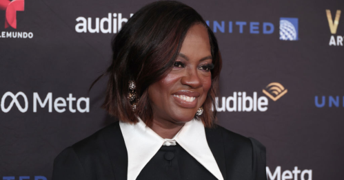Viola Davis smiles at a red carpet event, wearing a black outfit with a white collar, against a branded backdrop.