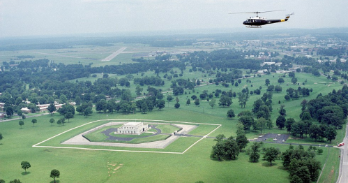Aerial view of a helicopter flying over a large park with a monument and surrounding greenery.