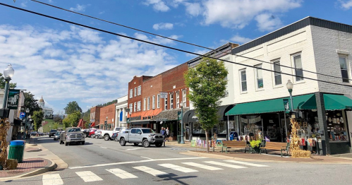 A charming small-town street with shops, trees, and a blue sky, featuring a crosswalk and parked cars.