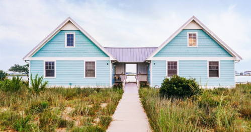 A charming blue beach house with a pathway, surrounded by grass and near the water.