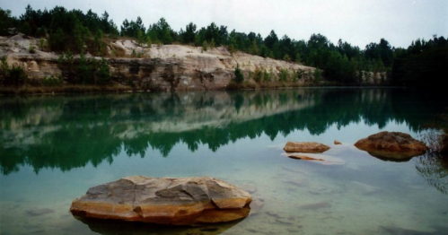 A tranquil lake surrounded by rocky cliffs and trees, with smooth stones visible in the water.