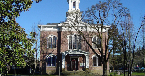 Historic brick building with a clock tower, surrounded by trees, housing the Jacksonville Museum. Clear blue sky.