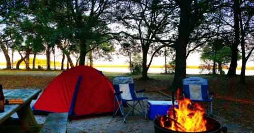 A cozy campsite with a red tent, two chairs, and a campfire surrounded by trees and a view of the water.