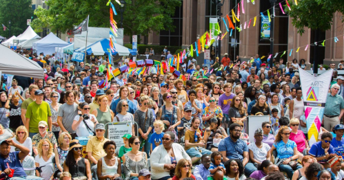 A large crowd gathers at an outdoor event, with colorful flags and tents in the background, enjoying the festivities.