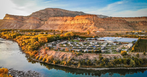 Aerial view of a campground by a river, surrounded by mountains and autumn-colored trees. RVs are parked in a circular layout.