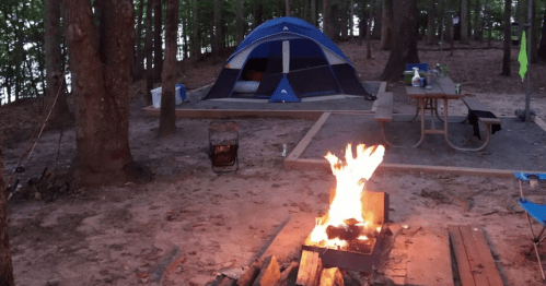 A campsite with a blue tent, a campfire, and a picnic table surrounded by trees near a lake.