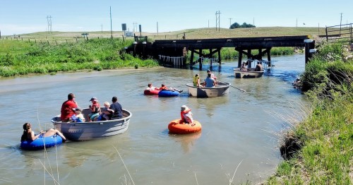 People in small boats and inner tubes float down a calm river, surrounded by green grass and a wooden bridge.