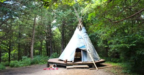 A teepee surrounded by trees, with a wooden platform and a blanket on the ground nearby.