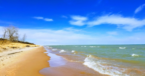 A serene beach scene with gentle waves, sandy shore, and a clear blue sky with scattered clouds.