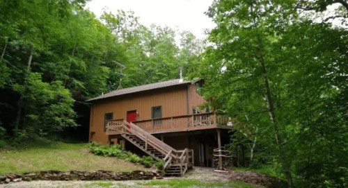 A wooden house surrounded by lush green trees, featuring a deck and stairs leading to the entrance.