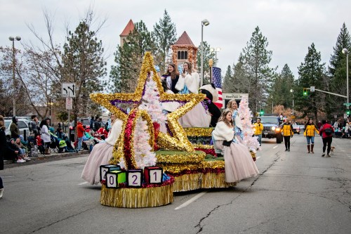 A festive parade float with women in elegant dresses, decorated with stars and trees, moving down a city street.