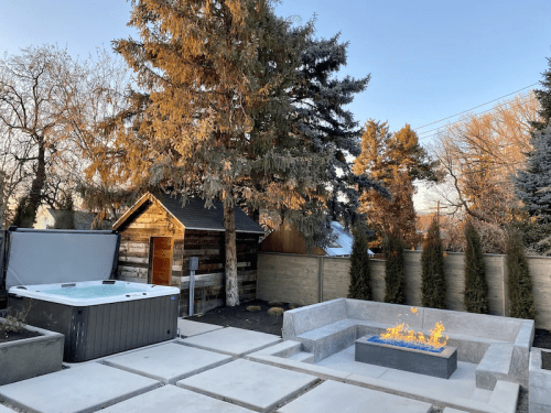 A backyard scene featuring a hot tub, fire pit, and a wooden shed surrounded by trees and a stone patio.