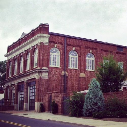 Historic brick building with arched windows and decorative details, surrounded by greenery and a cloudy sky.