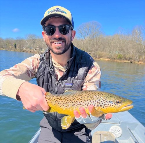 A smiling man in sunglasses holds a large brown trout while fishing on a sunny day.