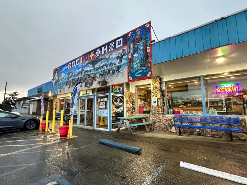 Colorful restaurant exterior with murals, outdoor seating, and a rainy atmosphere. Signs indicate it's open for business.