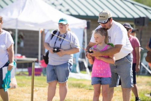 A man teaches a young girl to fish while others watch in a sunny outdoor setting.