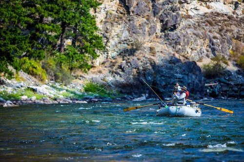 Two people fishing from a small raft on a river, surrounded by rocky cliffs and greenery.