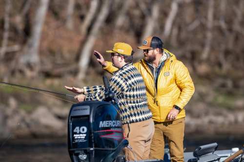 Two men fishing from a boat, one casting a line while the other gestures, surrounded by trees in the background.