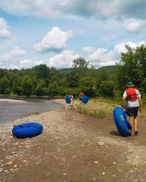 Three people walk along a riverbank carrying blue inner tubes, with trees and clouds in the background.