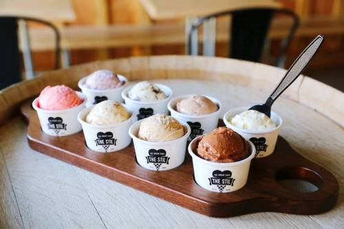 A wooden tray holds eight cups of various ice cream flavors, each labeled with a logo. A spoon rests in one cup.