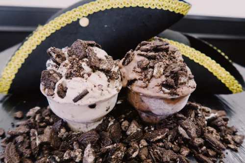 Two ice cream cups topped with crushed cookies, surrounded by cookie crumbs, with pirate hats in the background.