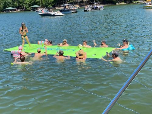 A group of people enjoying a sunny day on a floating mat in a lake, surrounded by boats and greenery.