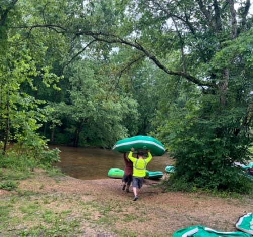 A person in a bright shirt carries a green kayak towards a river, surrounded by lush greenery.