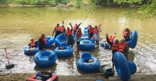 A group of people in blue inner tubes on a river, wearing life jackets and waving happily.