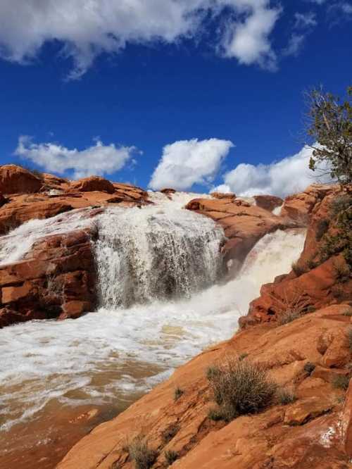 A waterfall cascades over red rock formations under a blue sky with fluffy white clouds.