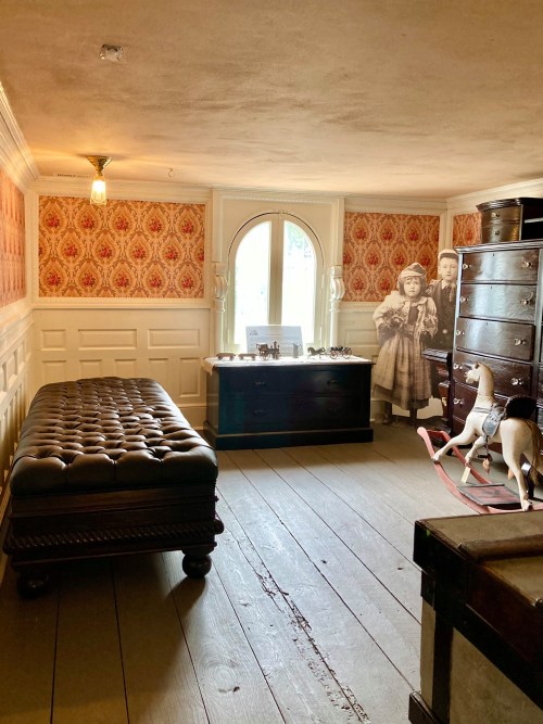 A vintage room with patterned wallpaper, a tufted bench, a mirror, and a rocking horse, featuring historical photographs.