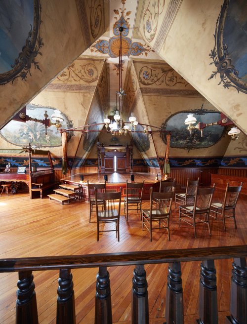 A spacious attic with a wooden floor, featuring ornate murals, vintage lamps, and a circle of chairs for gatherings.