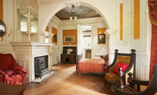 A vintage room with ornate decor, featuring a fireplace, a bed, and elegant seating, illuminated by natural light.
