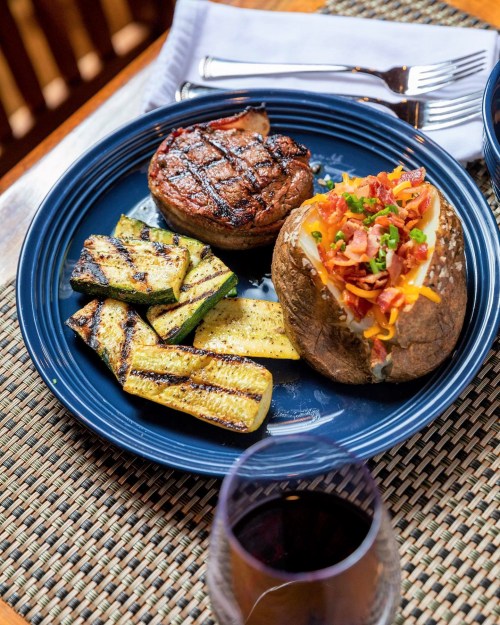A plate with a grilled steak, baked potato topped with cheese and bacon, and grilled zucchini, alongside a glass of red wine.