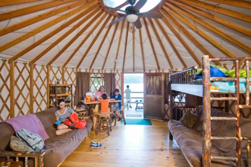 Interior of a yurt with wooden furniture, a bunk bed, and people relaxing and socializing. Natural light from the entrance.