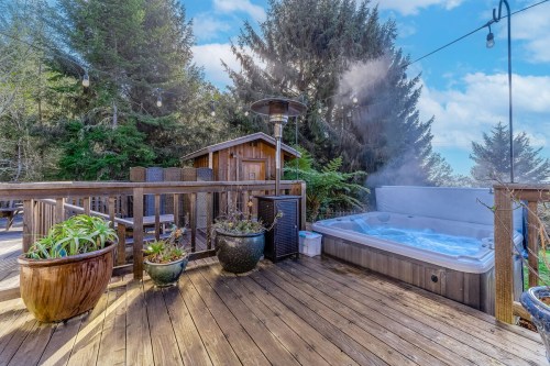 A wooden deck with a hot tub, surrounded by greenery and trees, featuring potted plants and outdoor lighting.