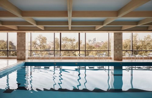 Indoor swimming pool with large windows overlooking trees and a cityscape, featuring a calm, reflective water surface.