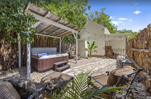 A cozy outdoor space featuring a hot tub, seating area, and lush greenery, with a white building in the background.