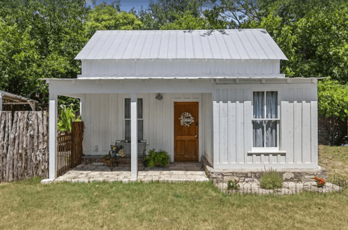 A small white cottage with a metal roof, wooden door, and a front porch surrounded by greenery and a stone pathway.