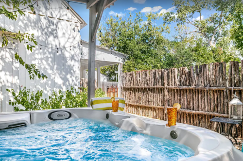 A hot tub with two drinks sits under a shaded patio, surrounded by greenery and a rustic wooden fence.