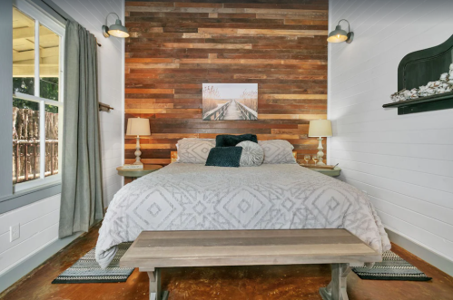Cozy bedroom with a wooden accent wall, a large bed, two lamps, and a bench at the foot. Natural light from a window.