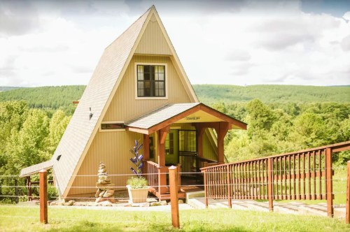 A charming A-frame house with a wooden porch, surrounded by lush greenery and rolling hills in the background.