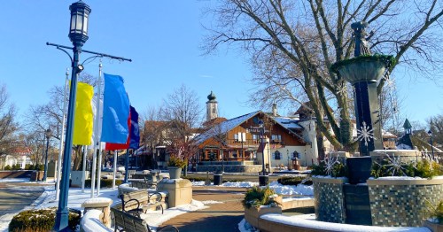 A winter scene of a town square with flags, benches, and a fountain, surrounded by snow and clear blue skies.