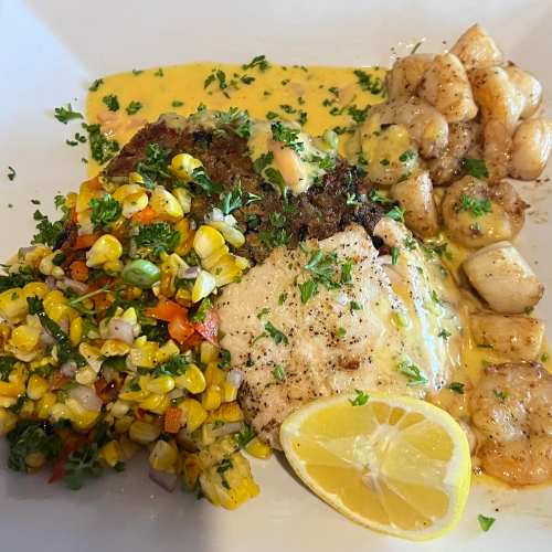 A plate featuring grilled fish with lemon, corn salad, and sautéed gnocchi in a creamy sauce, garnished with herbs.